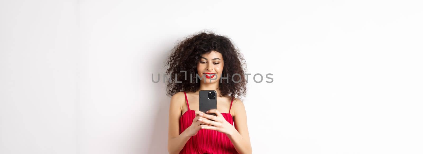 Happy fashionable woman taking photo on smartphone, photographing something on mobile and smiling, standing in red dress on white background.