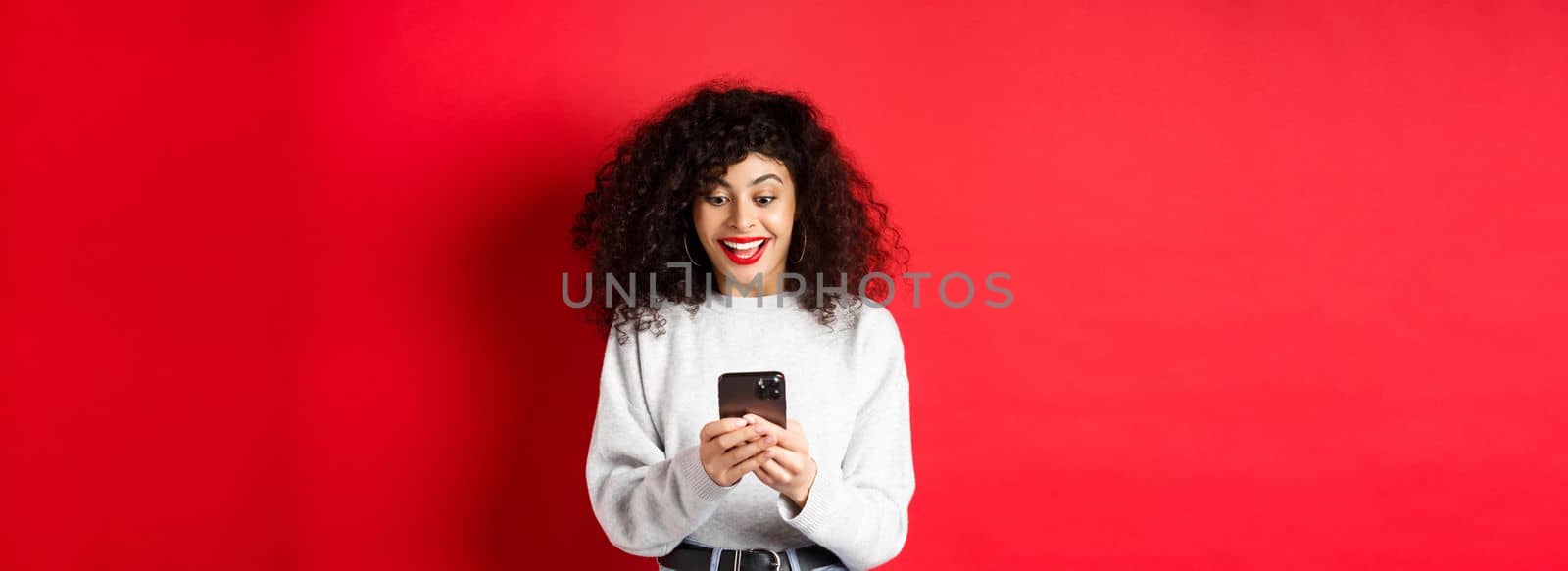 Image of young woman with curly hair, reading message on smartphone and smiling happy, receiving good news online, standing on red background.