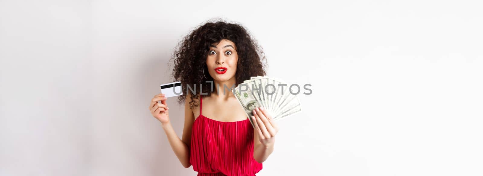 Excited woman looking with amazement and disbelief, showing dollars prize and plastic credit card, standing in red dress on white background.