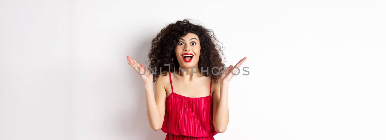 Surprised happy woman reacting to big news, looking cheerful with hands spread sidewas, standing in red dress on white background.