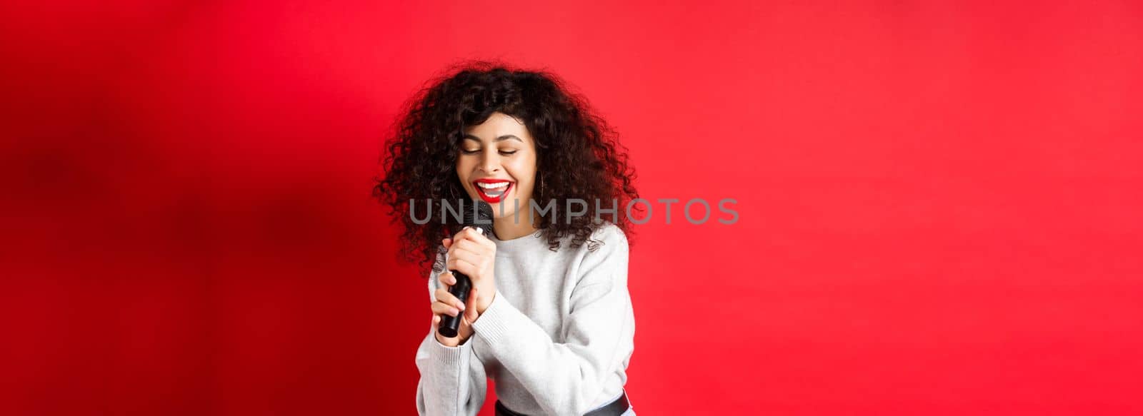 Hobbies and leisure concept. Beautiful woman singing in microphone and smiling, having fun at karaoke, standing on red background.