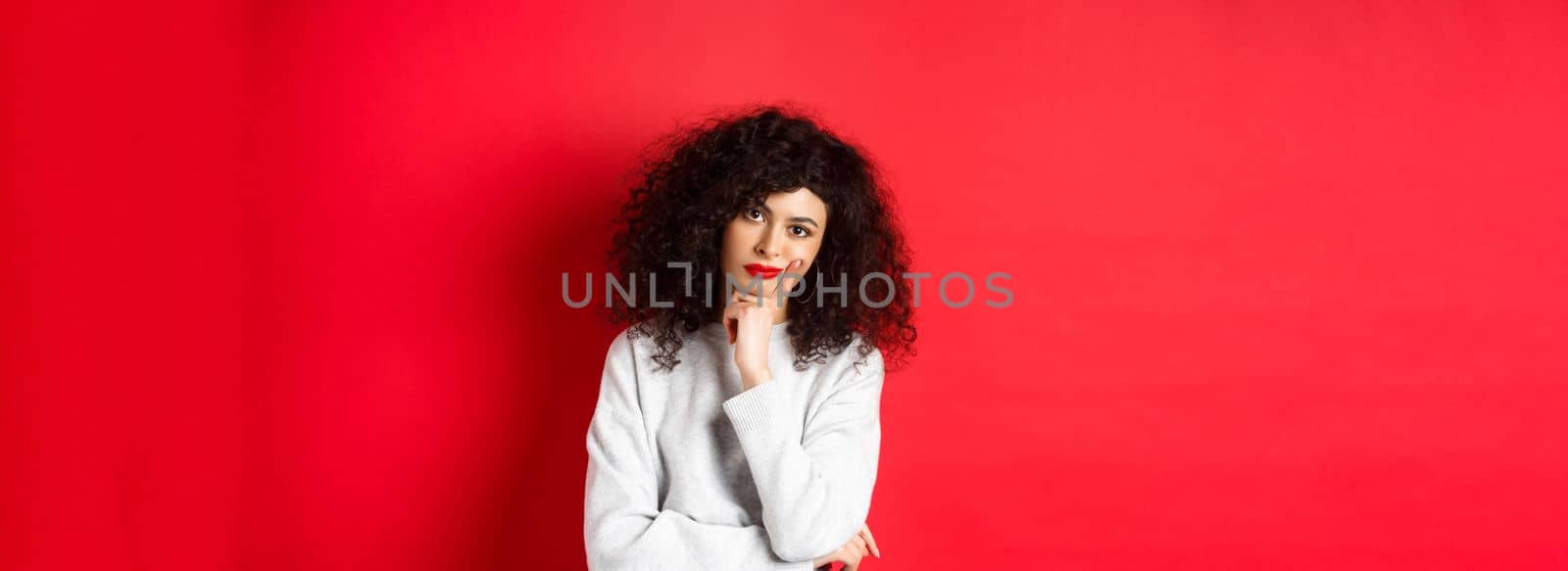 Bored and tired curly woman with red lips, looking bothered at camera, standing against studio background.