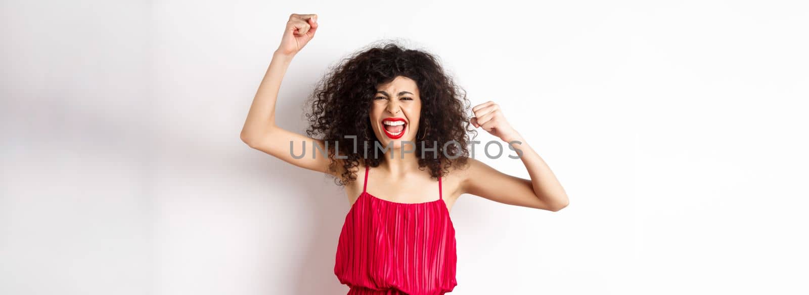 Cheerful emotive woman with curly hair, red dress, raising hands up and chanting, rooting for team, shouting wanting to win, standing on white background.