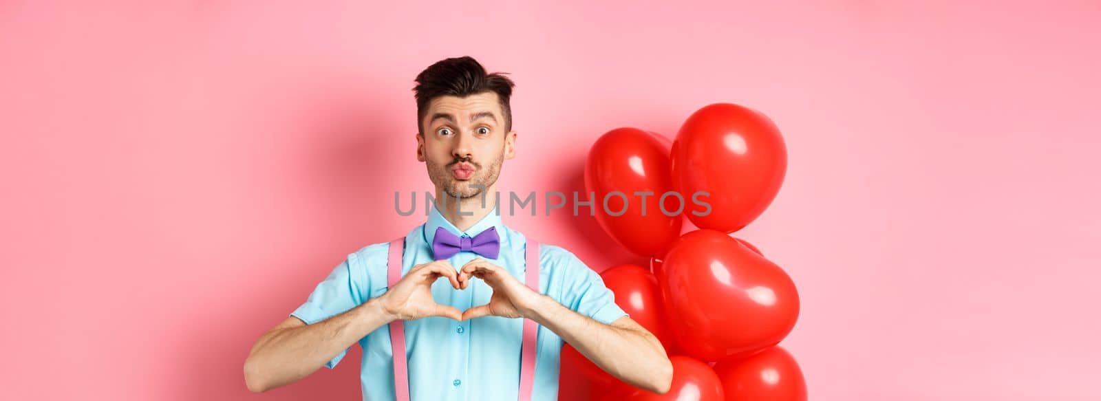 Valentines day concept. Cute boyfriend pucker lips for kiss, standing in bow-tie with heart gesture, say I love you, standing on pink background.