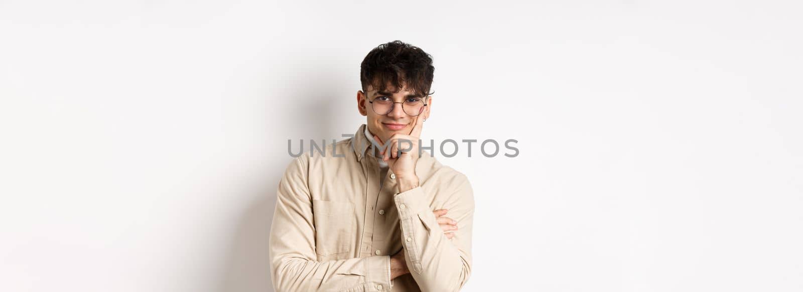 Cunning young man watching something interesting happen, lean face on arm and smiling devious, looking at camera intrigued, standing on white background by Benzoix