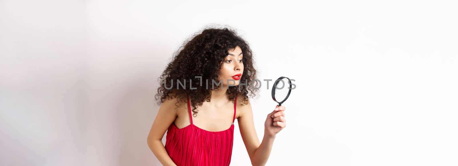 Beautiful woman in red dress searching for something, looking aside through magnifying glass, standing against white background by Benzoix