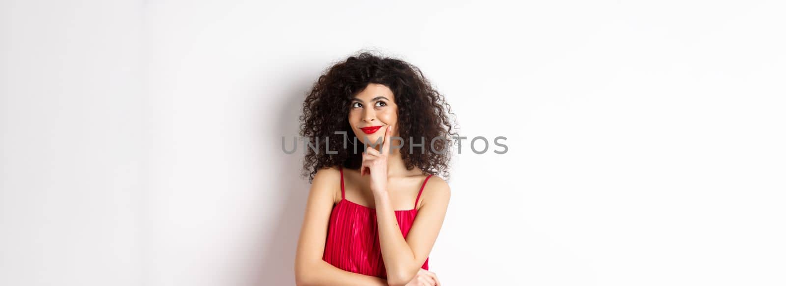 Image of elegant woman in red dress, looking pensive and smiling, thinking of romantic date, standing over white background.