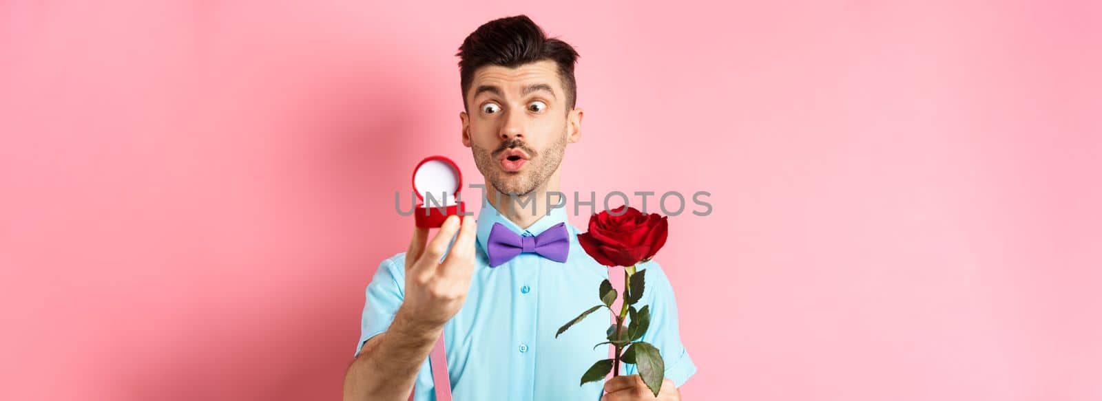 Valentines day. Romantic funny guy going to make wedding proposal, asking to marry him, holding red rose and looking at engagement ring with excitement, pink background.