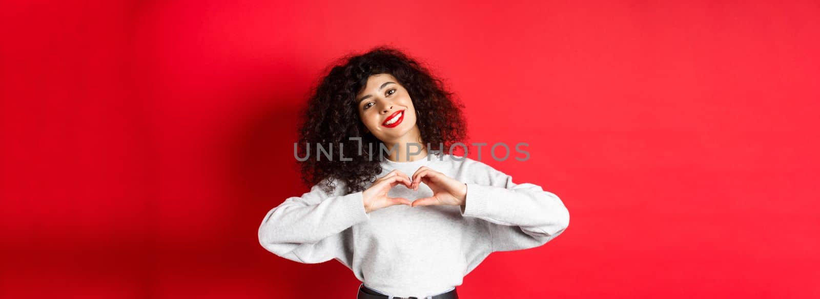 Beautiful young woman with curly hair showing heart gesture, say I love you and smile romantic at camera, standing on red background.