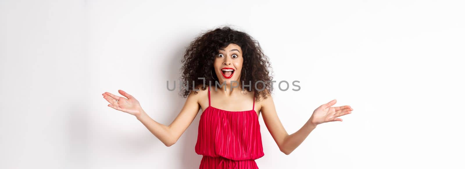 Excited young woman in red dress, spread hands sideways and screaming surprised, standing happy on white background.