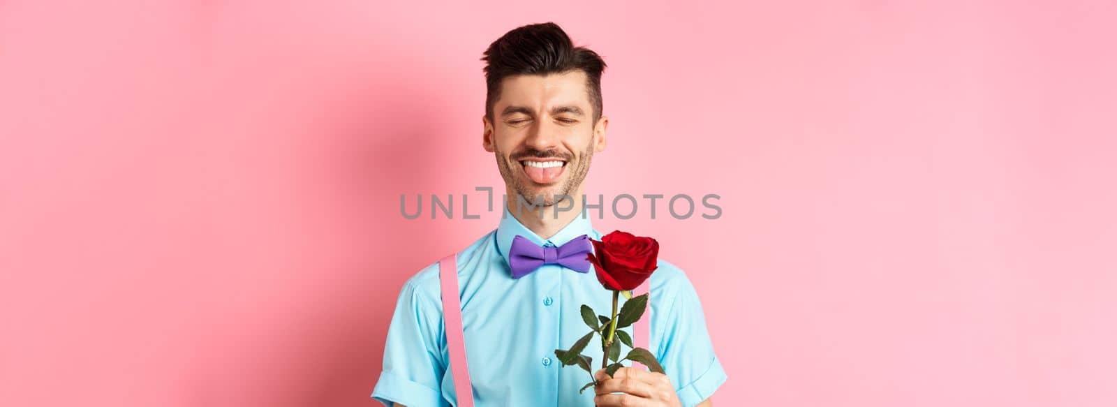 Happy man showing tongue and smiling, holding red rose for girlfriend on Valentines day, enjoying romantic date, pink background.