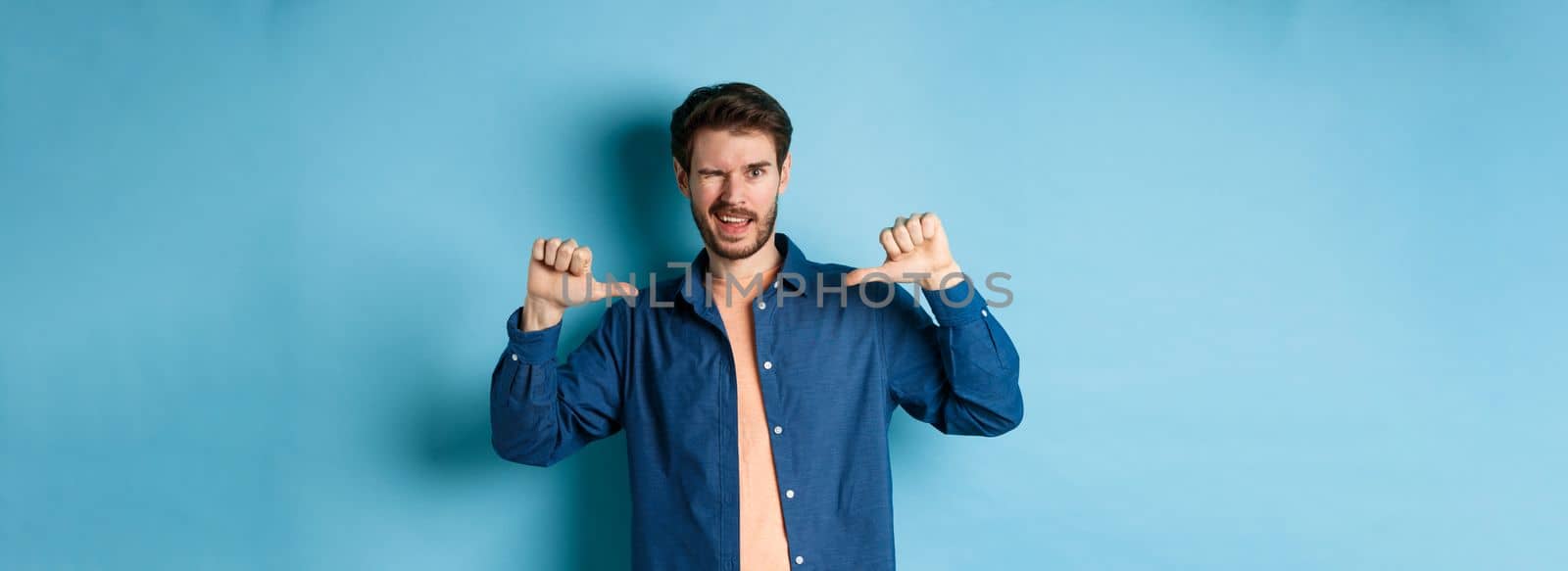 Confident man winking and pointing at himself, self-promoting, standing on blue background. Copy space