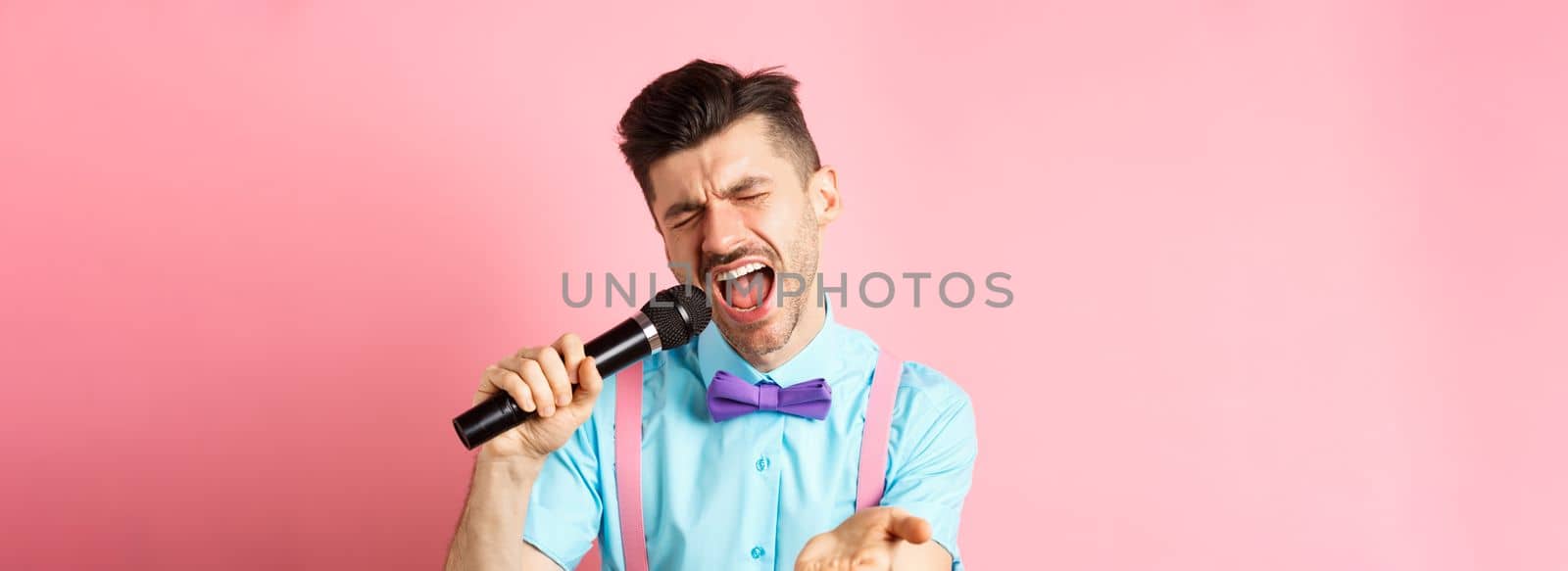 Party and festive events concept. Passionate singer holding microphone and pointing hand at you, singing about love, standing on pink background.