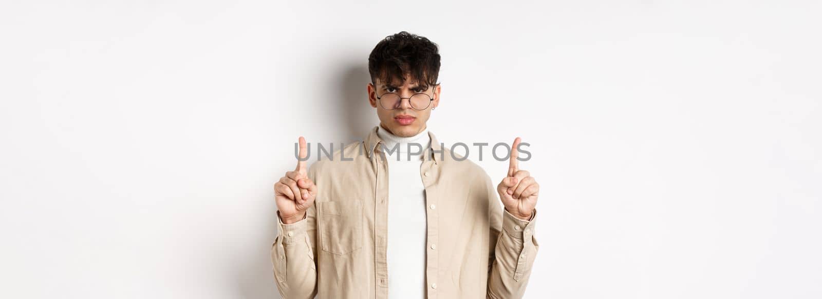 Angry and displeased young man in glasses frowning, looking disappointed and pointing fingers up, standing on white background.