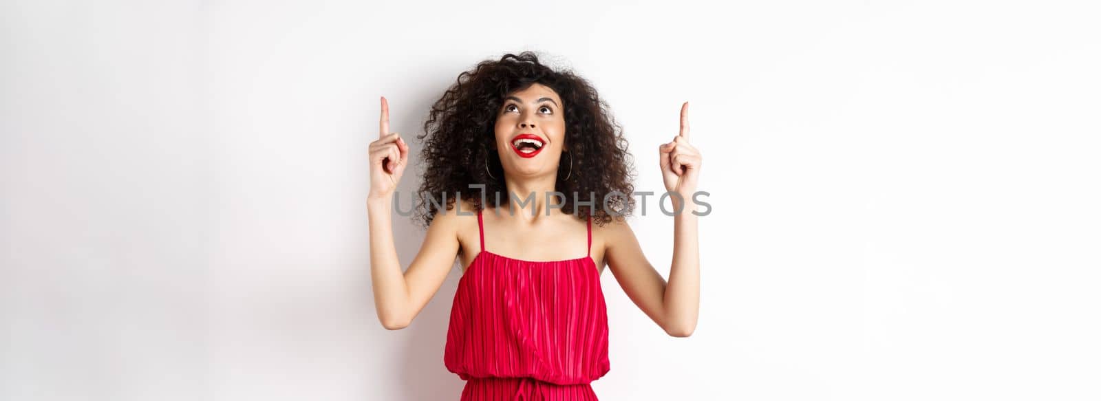 Excited caucasian woman with curly hair, red lips, looking and pointing up with amazed smile, checking out promo offer, white background by Benzoix