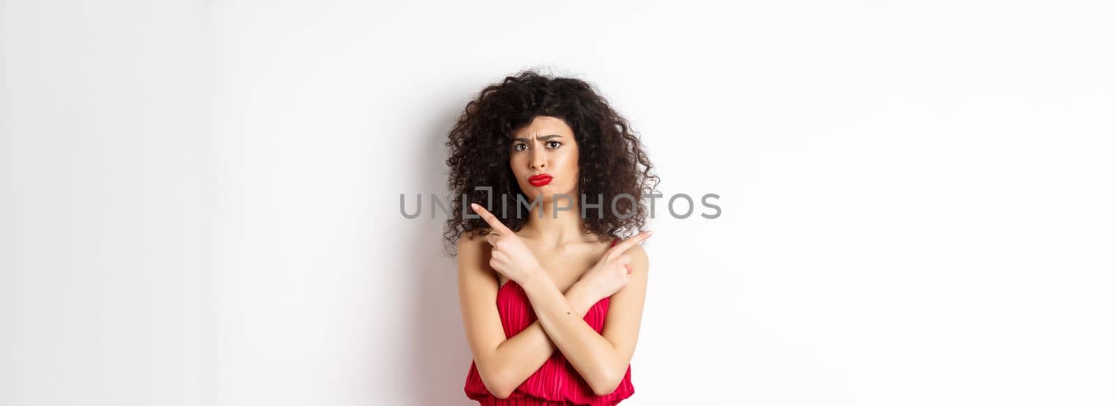 Doubtful and sad young woman in red dress, pointing fingers sideways and sulking, cant decide between products, need help with choice, standing over white background.