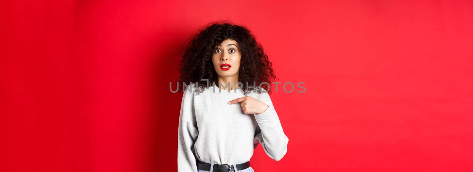 Surprised attractive female model pointing at herself, raising eyebrows and looking with disbelief at camera, standing on red background.