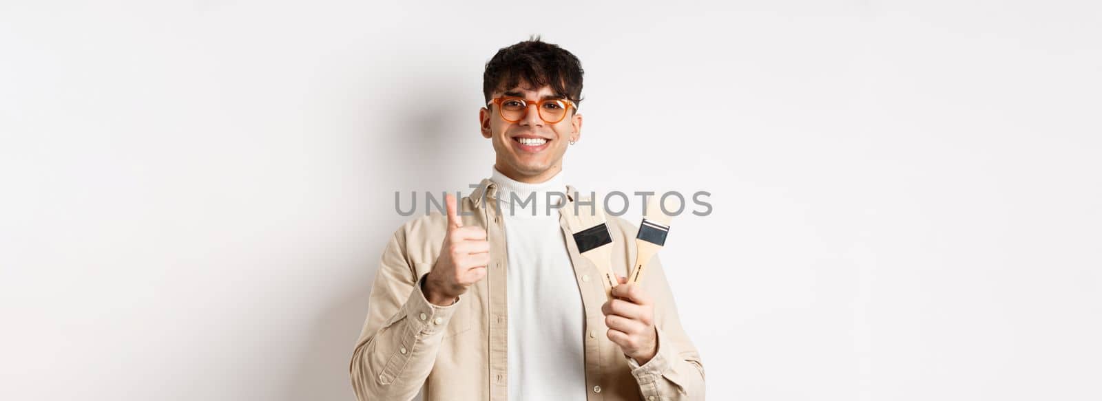 Handsome natural guy in glasses recommending store, showing painting brushes for renovation and decor, make thumbs up in approval, standing on white background.