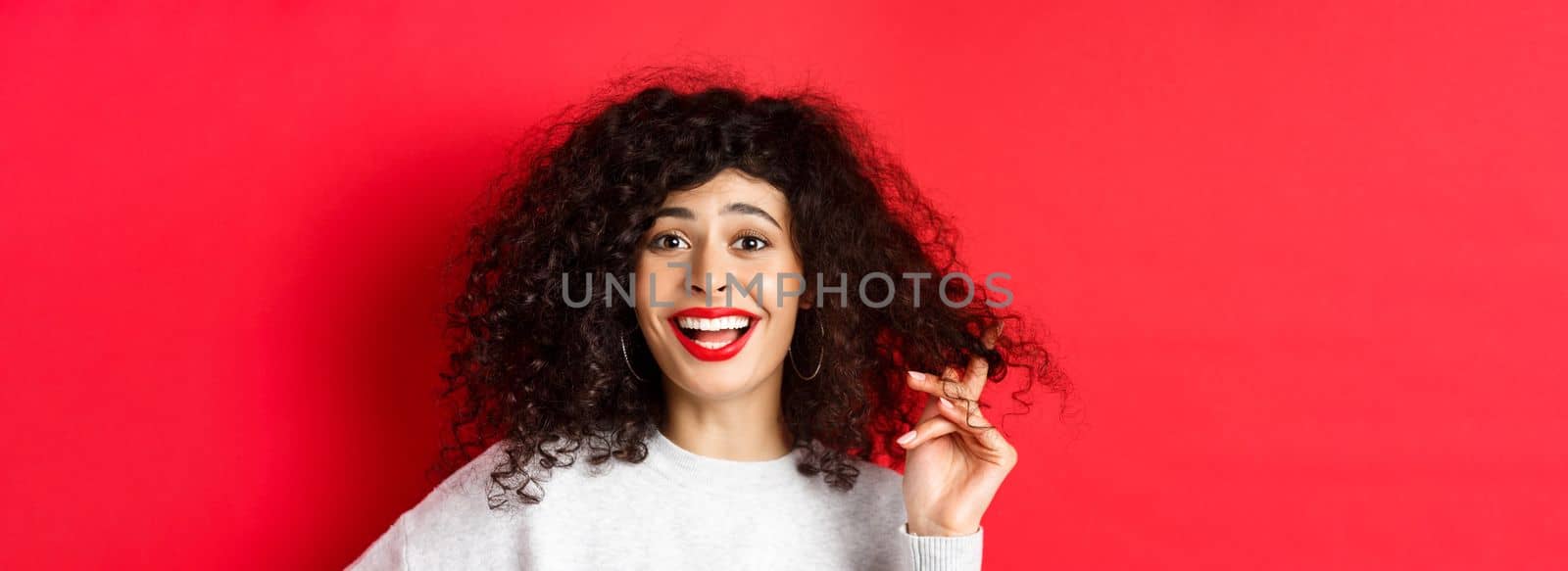 Close-up of happy woman playing with curly hair and laughing at something funny, standing on red background by Benzoix