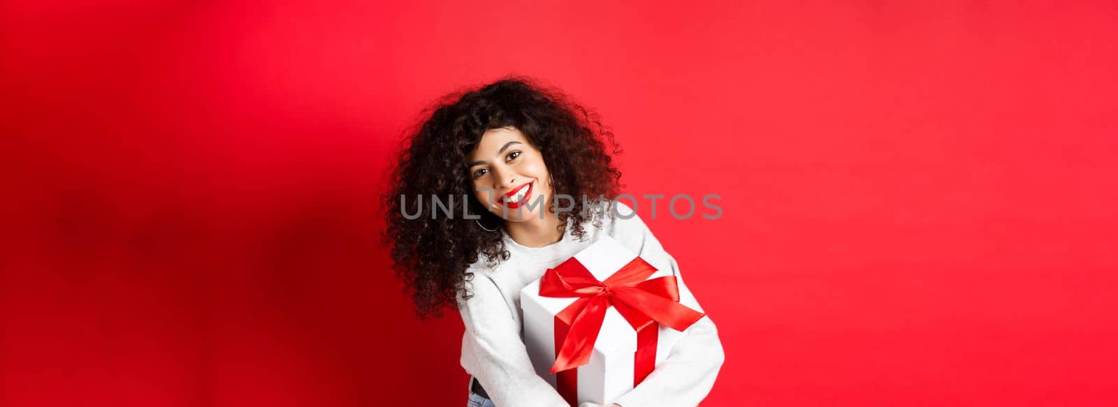 Celebration and holidays concept. Happy woman holding birthday gift and smiling at camera, standing in casual clothes, red background.