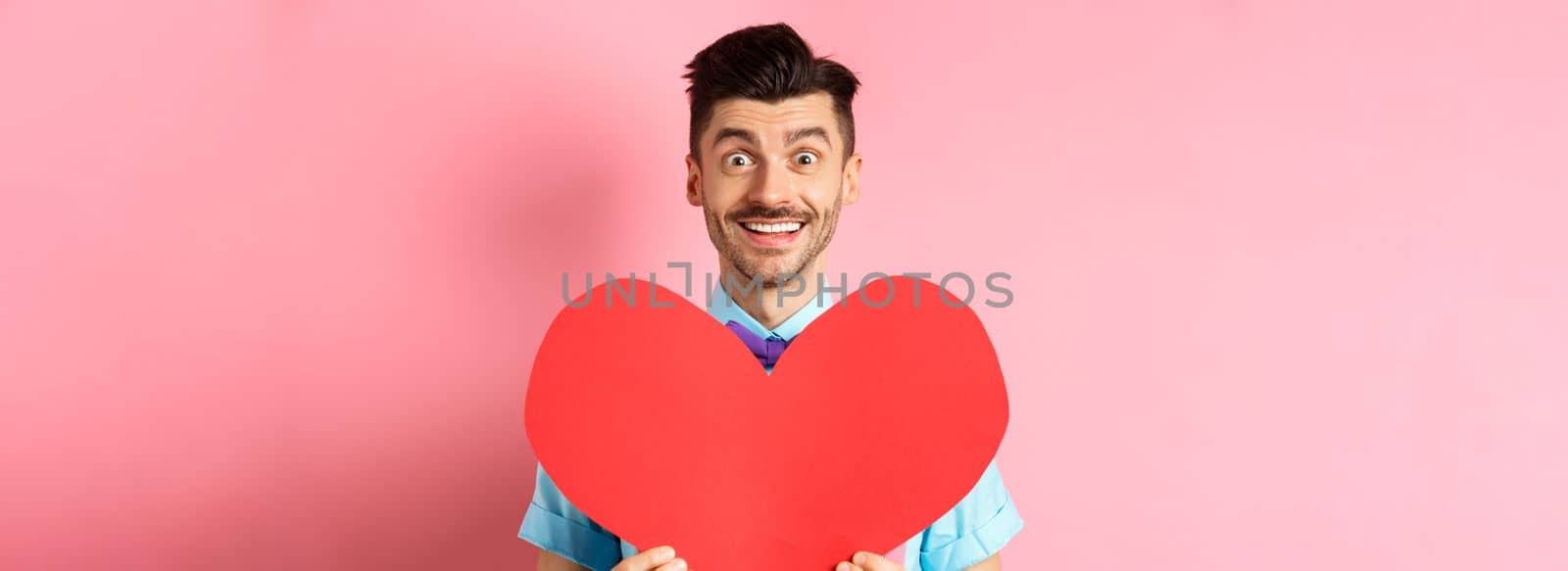 Hopeful man in love showing red heart sign, smiling at camera, waiting for soulmate on Valentines day, standing on pink background.