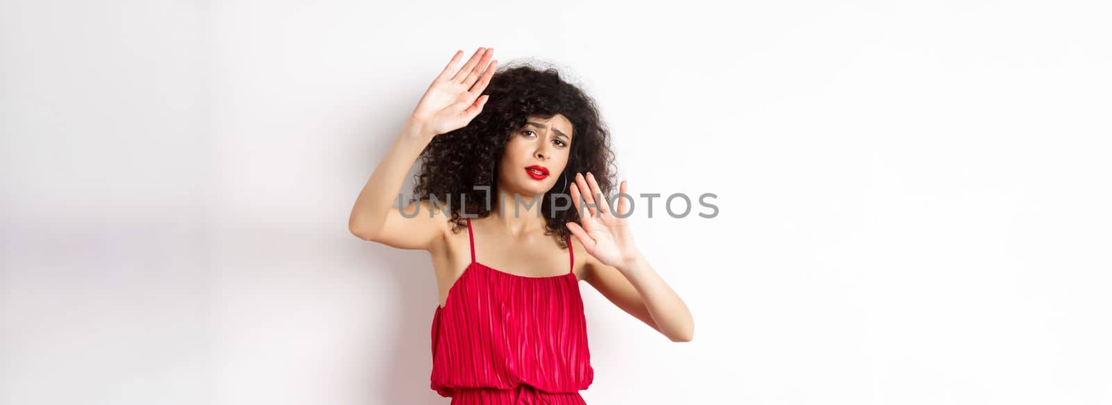 Young woman with curly hair and red dress, asking to stop, block someone, raising hands defensive, protecting herself, standing against white background by Benzoix