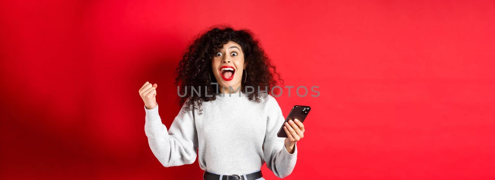 Excited caucasian woman with curly hair, chanting, winning online prize, holding smartphone and triumphing, standing on red background by Benzoix