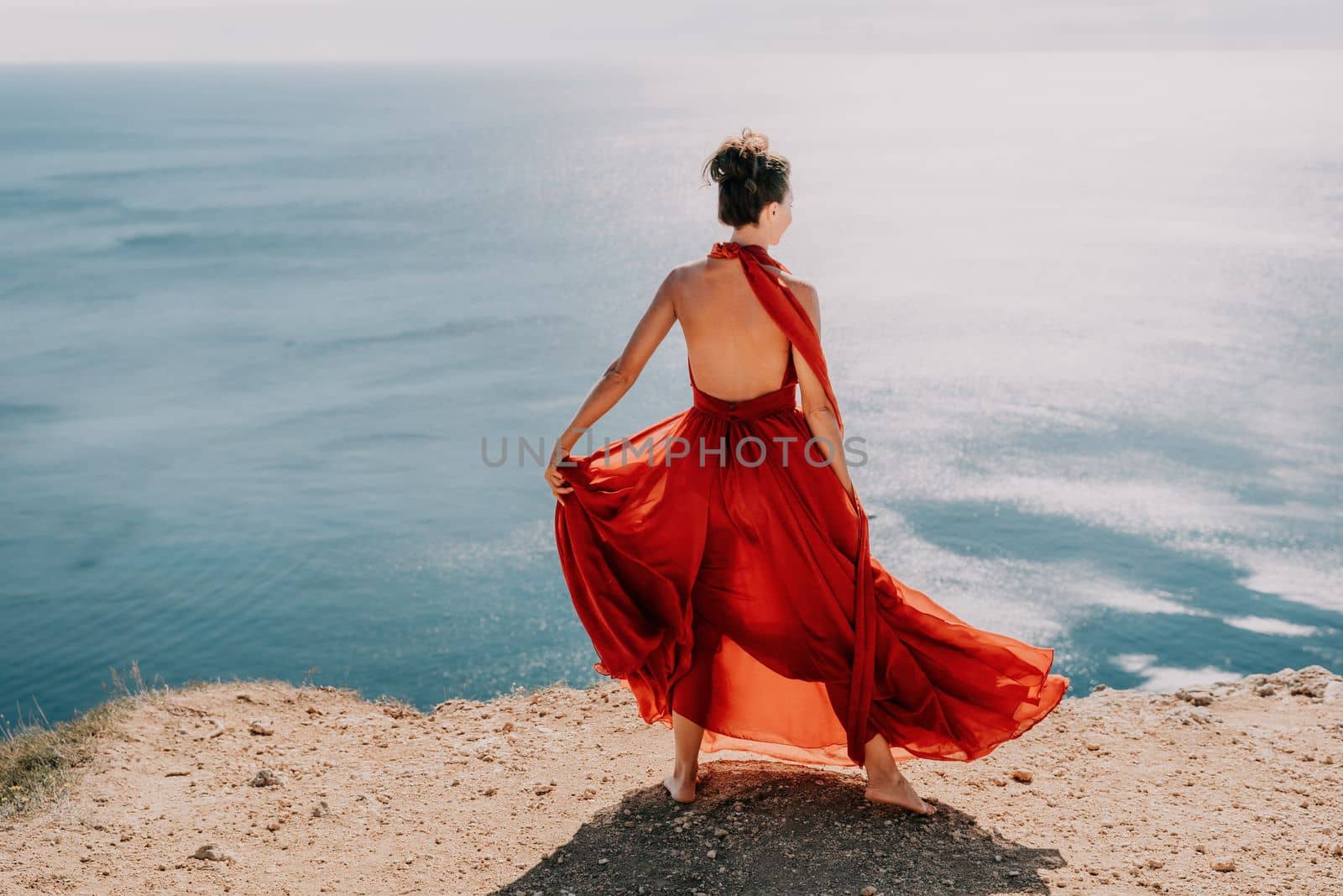 Side view a Young beautiful sensual woman in a red long dress posing on a rock high above the sea during sunrise. Girl on the nature on blue sky background. Fashion photo.