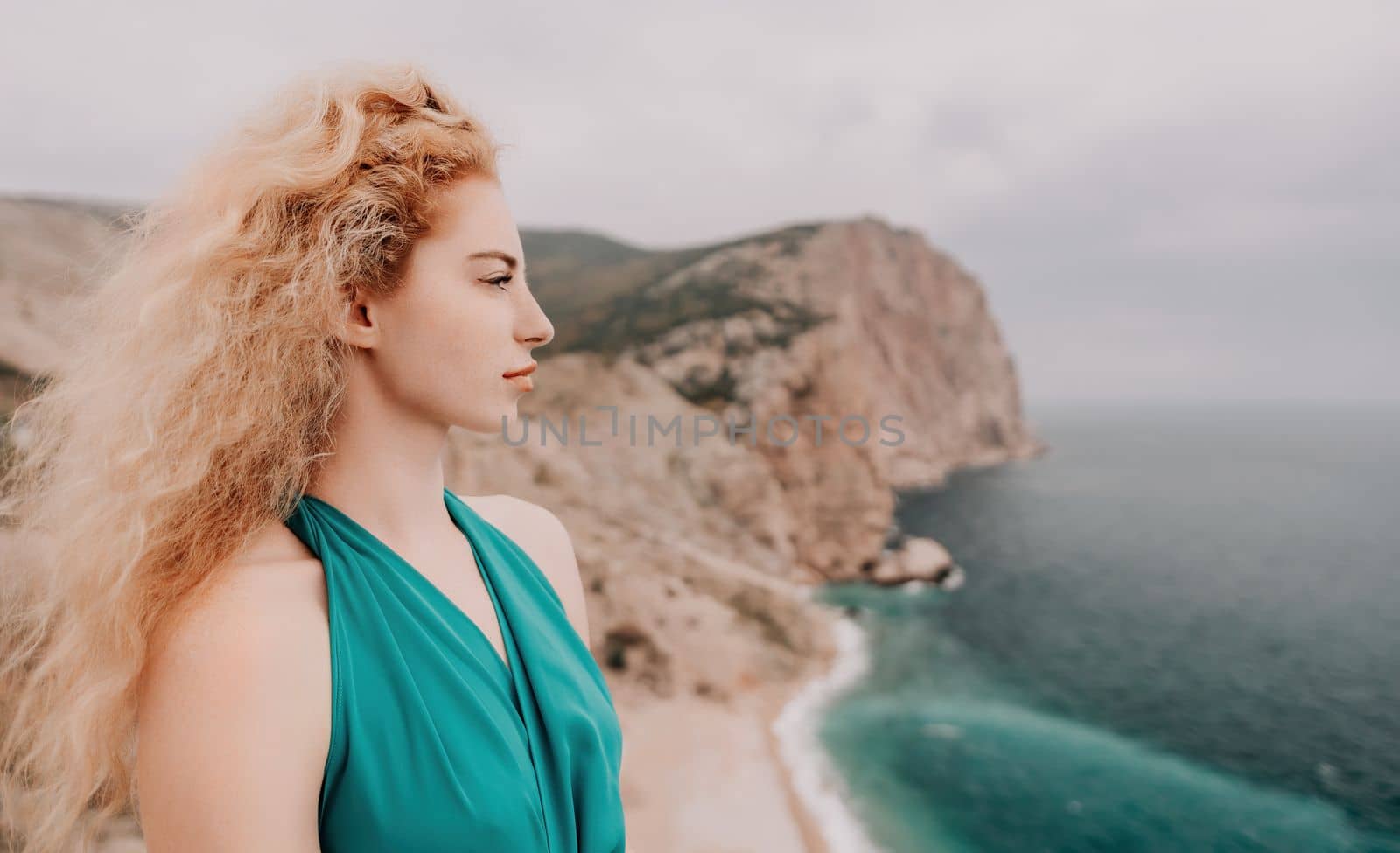 Side view a Young beautiful sensual woman in a mint long dress posing on a volcanic rock high above the sea during sunset. Girl on the nature on overcast sky background. Fashion photo