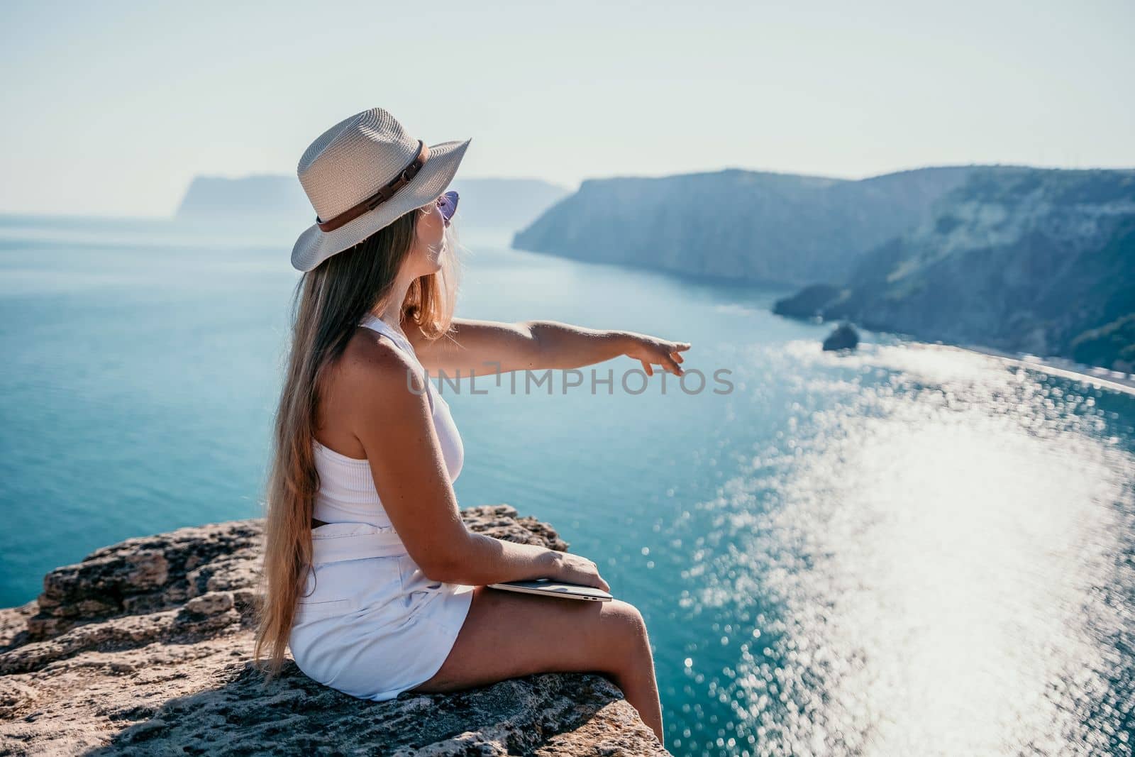 Successful business woman in yellow hat working on laptop by the sea. Pretty lady typing on computer at summer day outdoors. Freelance, travel and holidays concept.