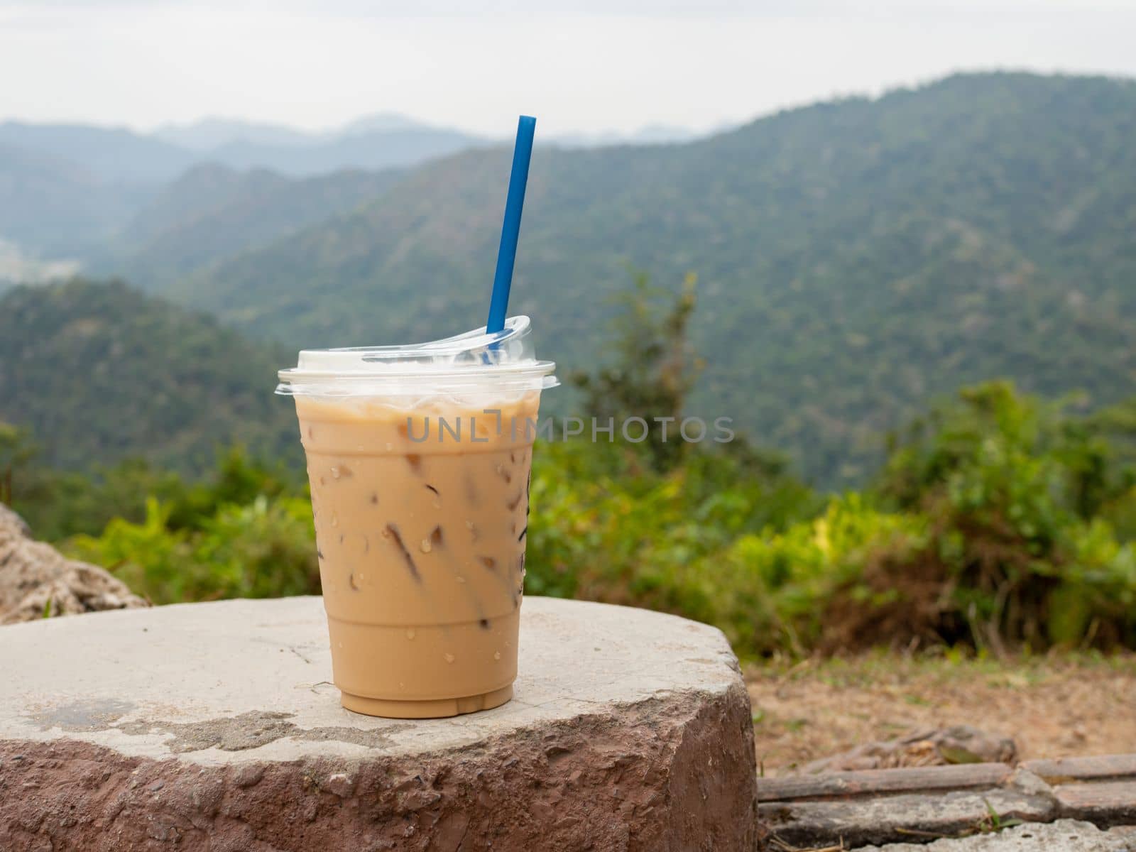 A glass of iced coffee is placed on a rock against a background of mountains and sky. by Unimages2527