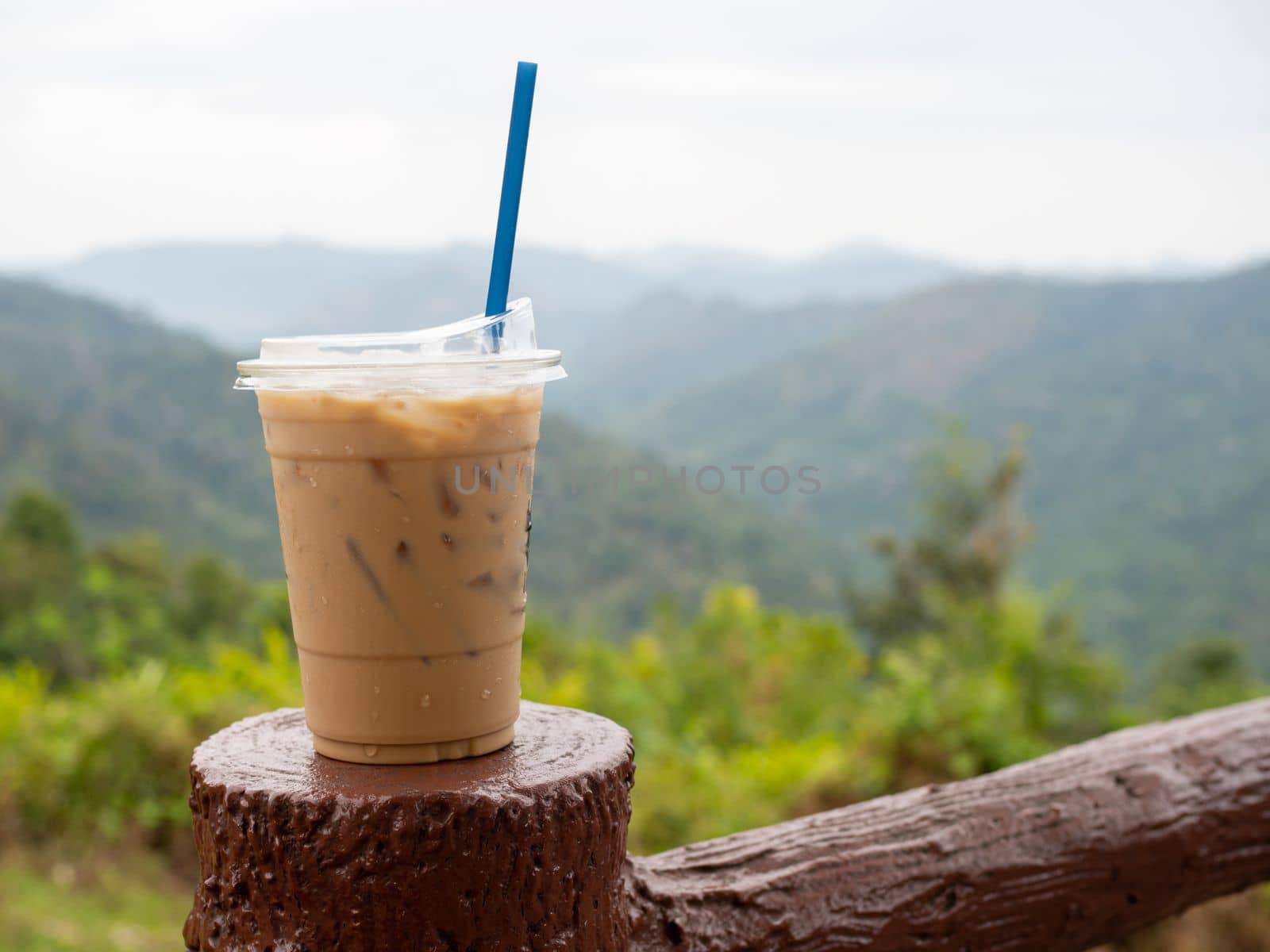 A glass of iced coffee is placed on the fence against a backdrop of mountains and sky. by Unimages2527