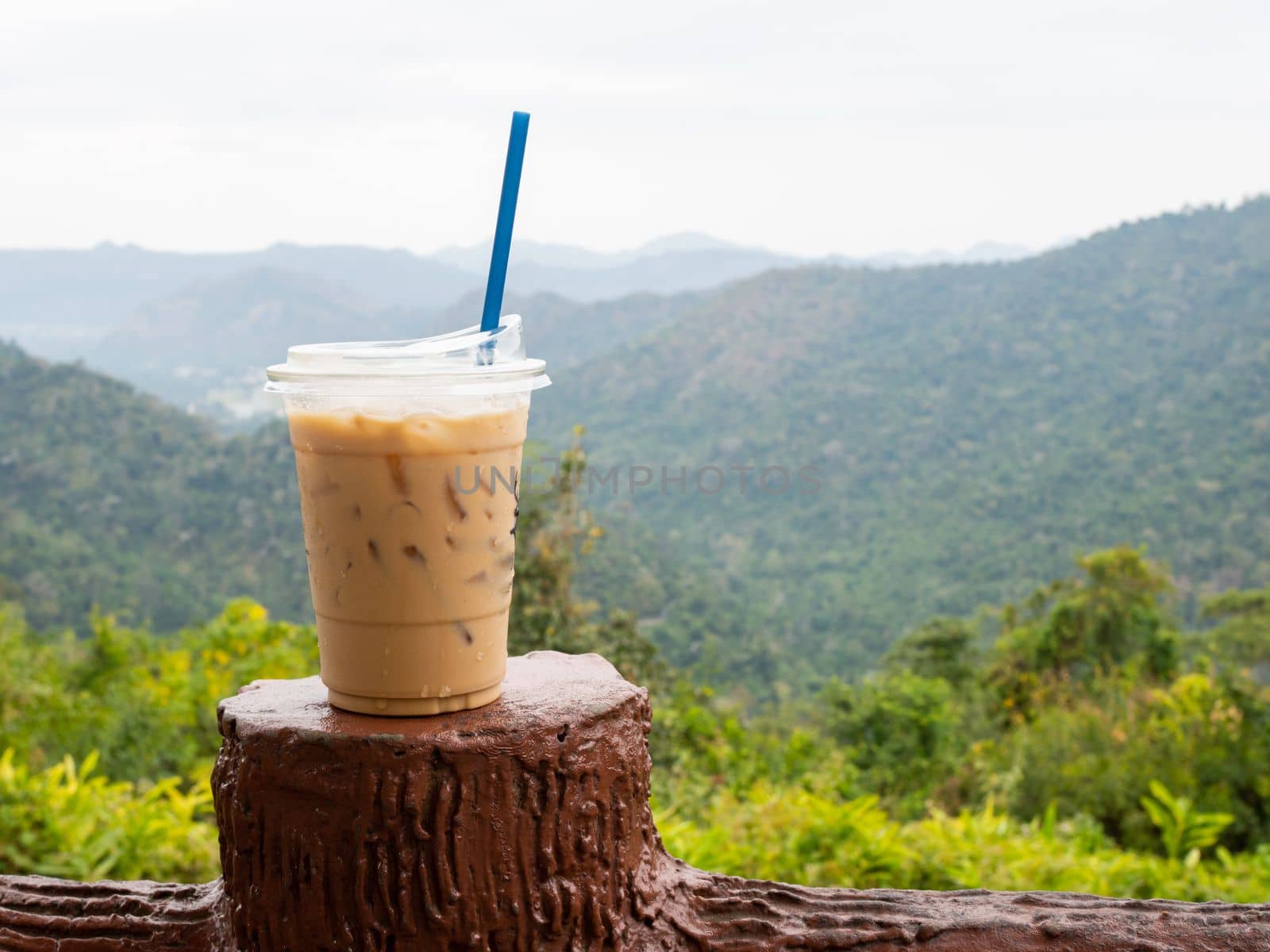 A glass of iced coffee is placed on the fence against a backdrop of mountains and sky. by Unimages2527