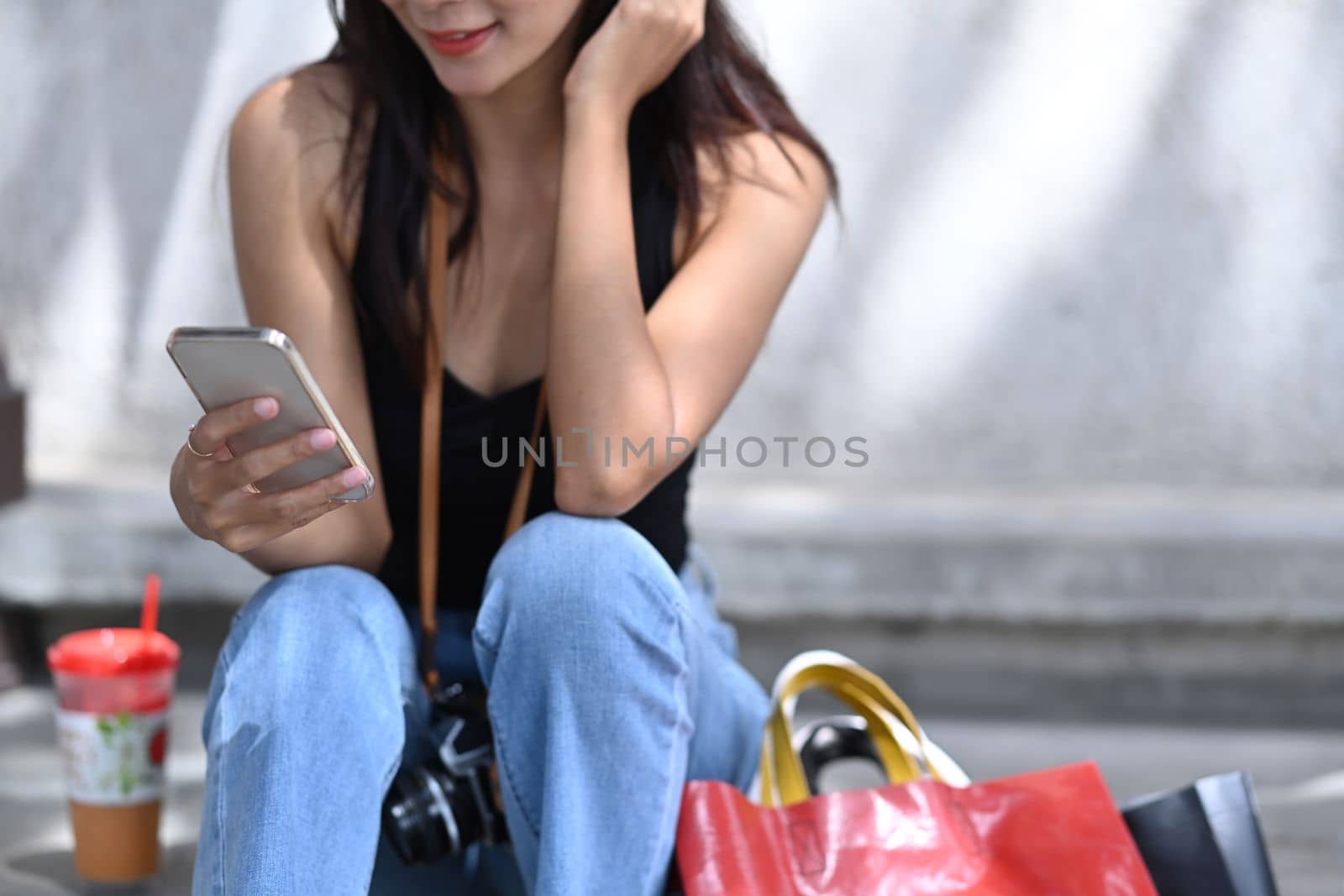 Smiling young woman using smart phone and sitting on stairs with shopping bags.