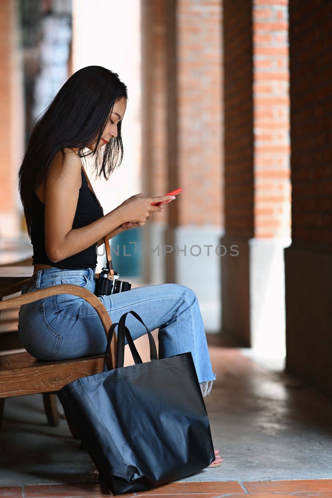 Full length portrait of beautiful woman sitting on bench and using smart phone.