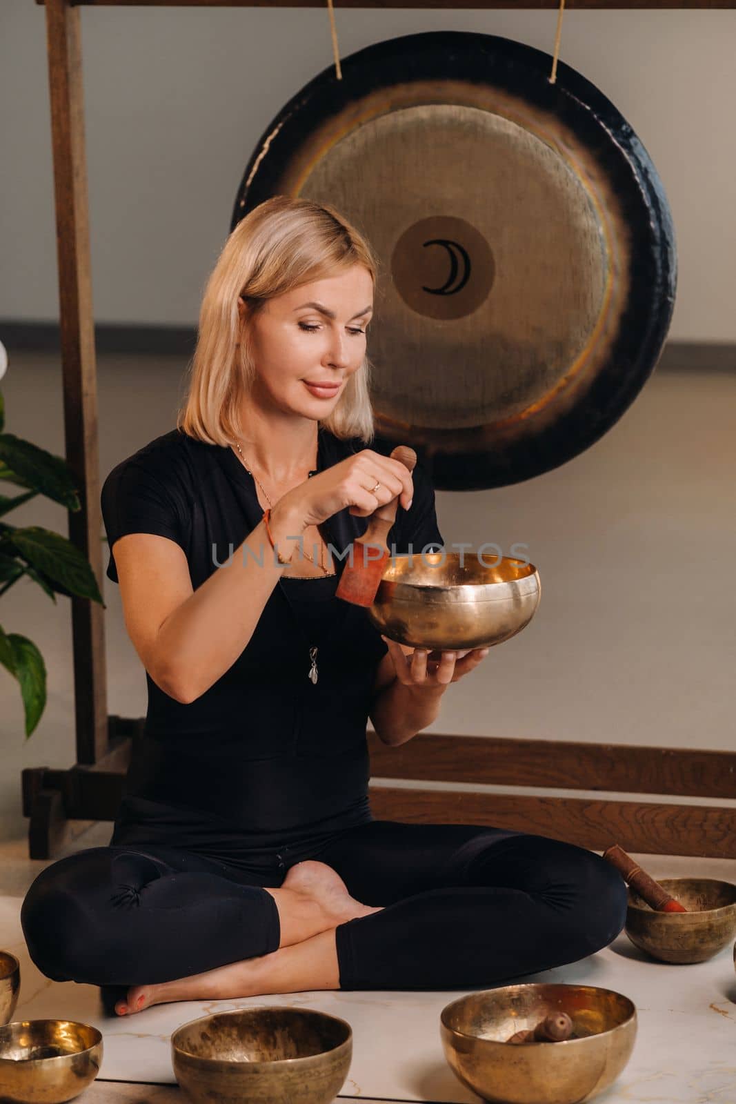 A woman plays a Tibetan singing bowl while sitting on a yoga mat against the background of a gong.