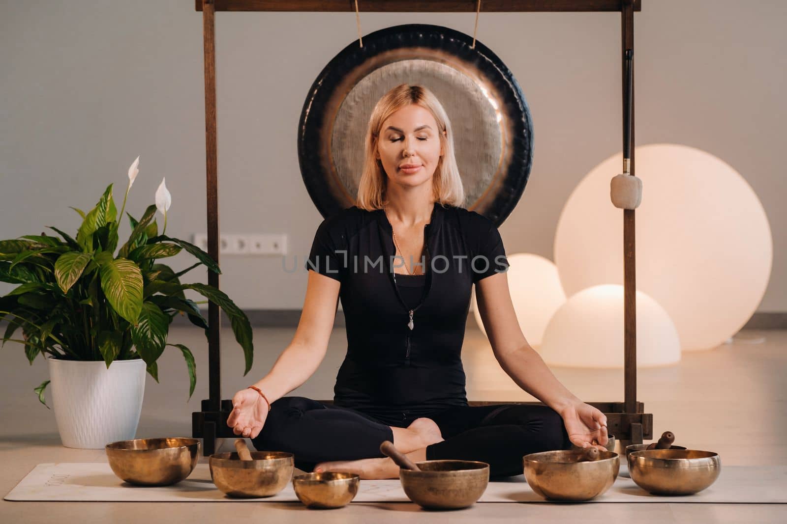 A woman sits in a lotus position next to Tibetan bowls, sitting on a yoga mat against the background of a gong by Lobachad