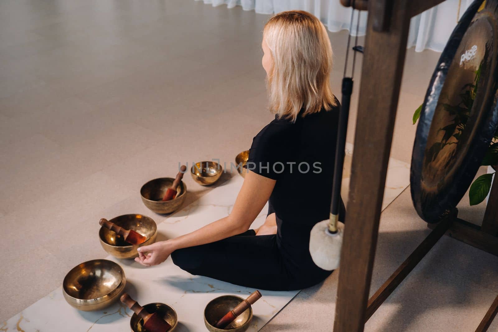 A woman sits in a lotus position next to Tibetan bowls, sitting on a yoga mat against the background of a gong.