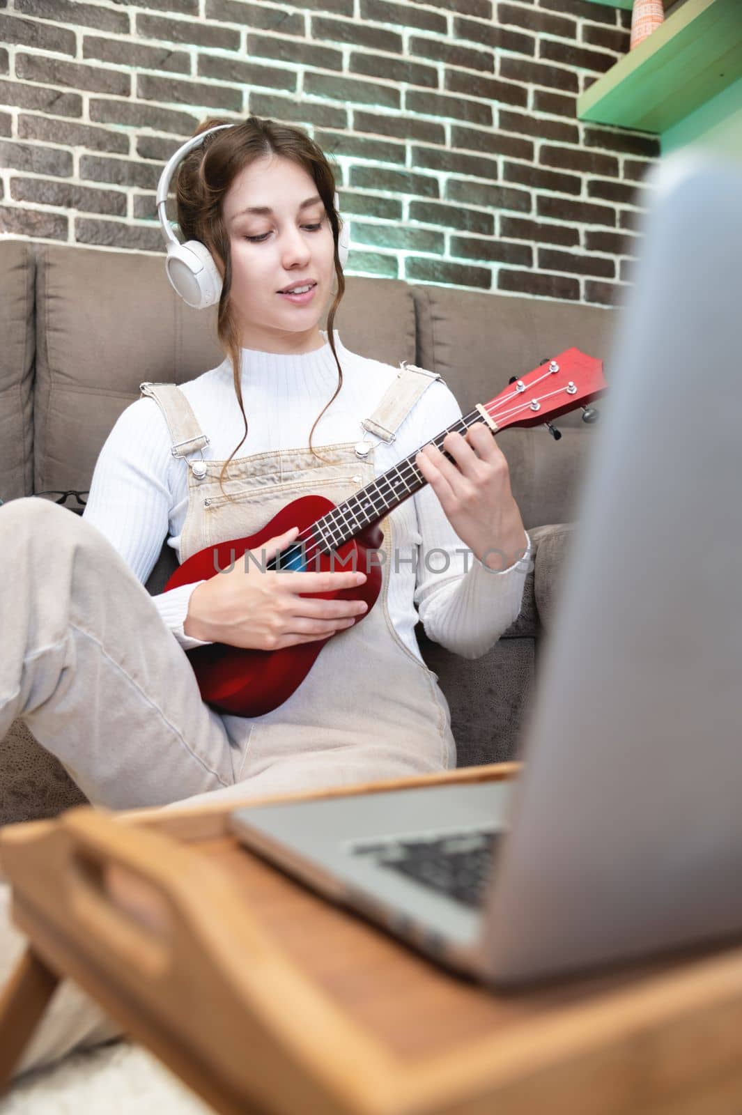 Smiling woman recording new content to vlog, playing on ukulele guitar, explains how to play, looking at webcam, sitting on couch at home during lockdown and quarantine. Online training, hobbies.