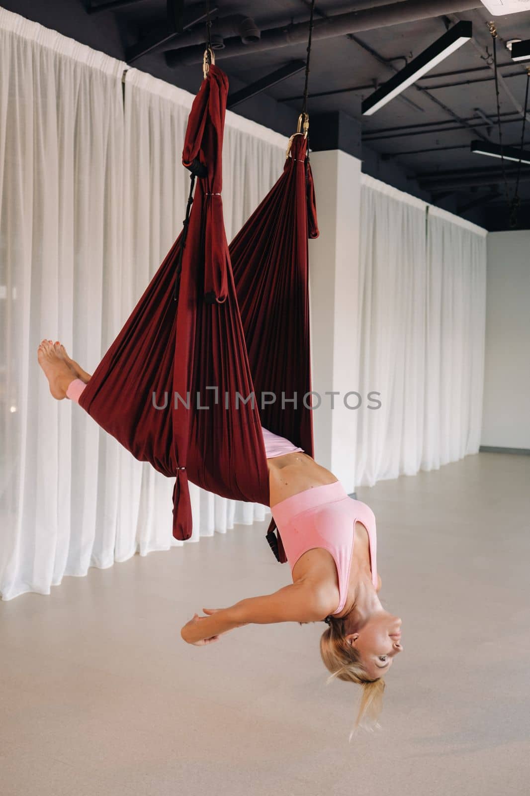 A young girl, an aero yoga coach, is hanging on a hammock in the yoga club hall. a woman in pink sportswear is doing anti-gravity yoga.