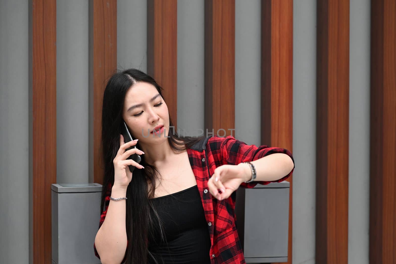 Young Asian woman talking on mobile phone and checking time on her wrist watch. by prathanchorruangsak