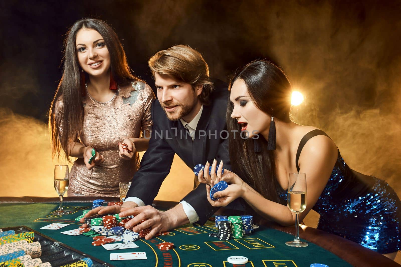 Two beautiful women and young man play on poker table in casino, focus on man and brunette by nazarovsergey