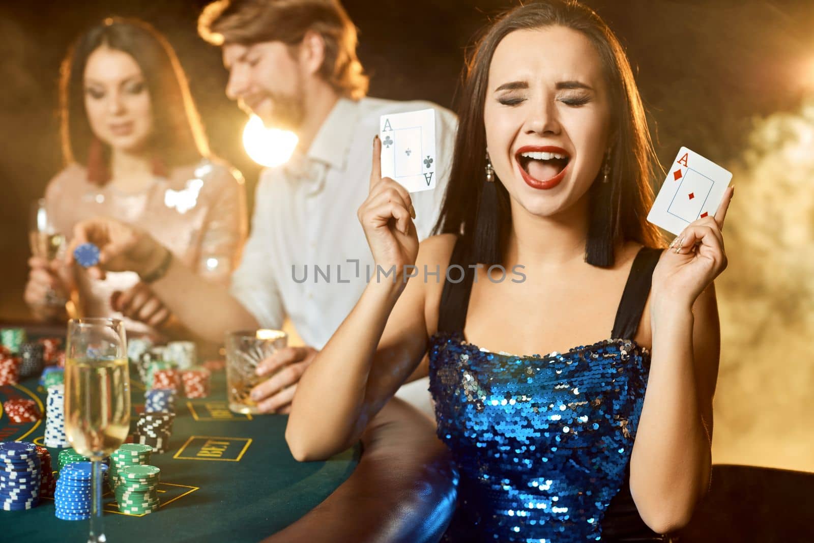Group of an elegant people playing poker at the gambling house. Focus on a emotional brunette in a blue shiny dress by nazarovsergey