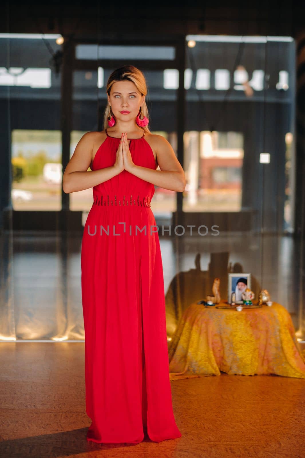 Portrait of a smiling girl in a red dress stands in the interior with her hands clasped in front of her.