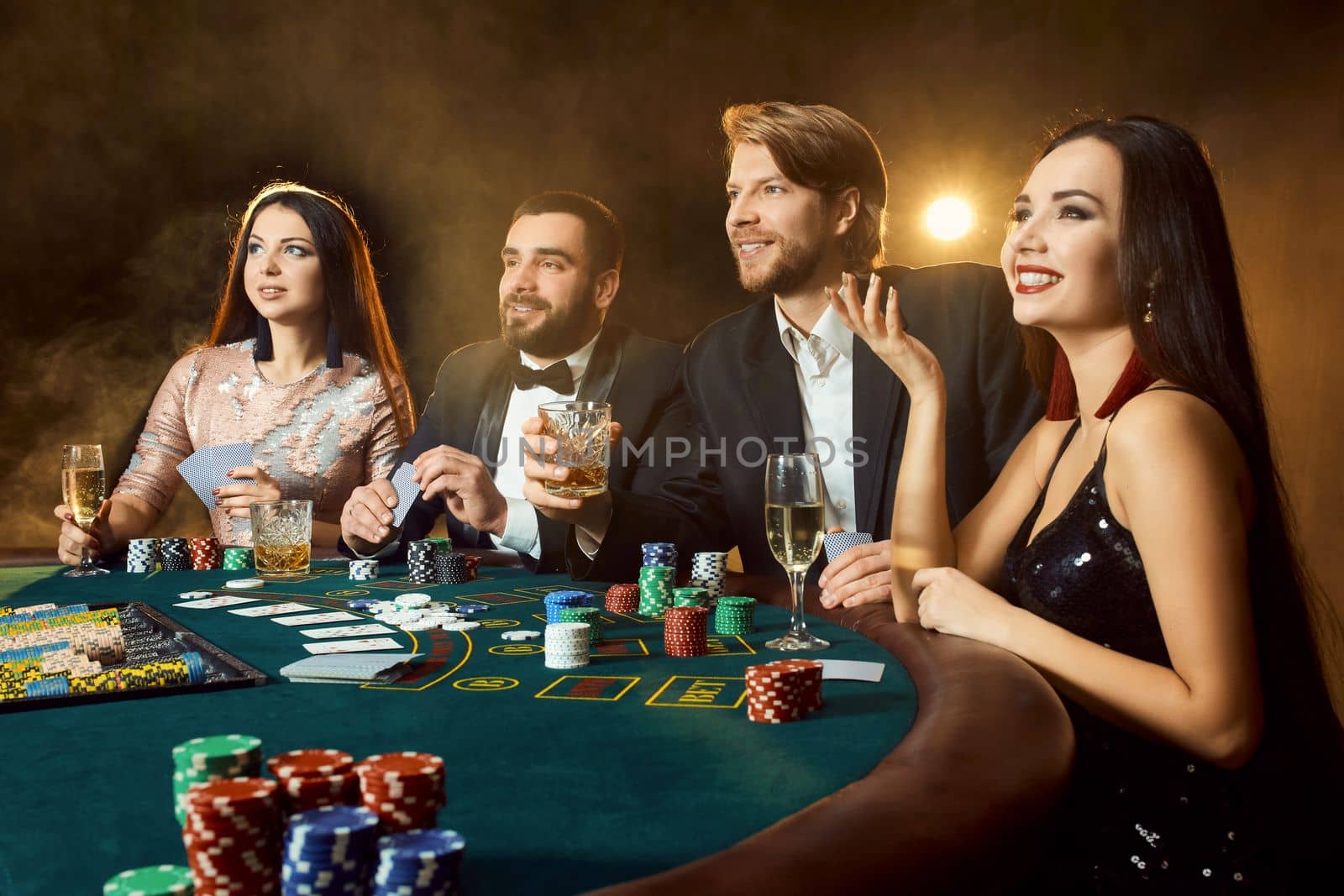 Upper class friends gambling in a casino. Two men in suits and two young women in dresses. Smoke. Casino. Poker