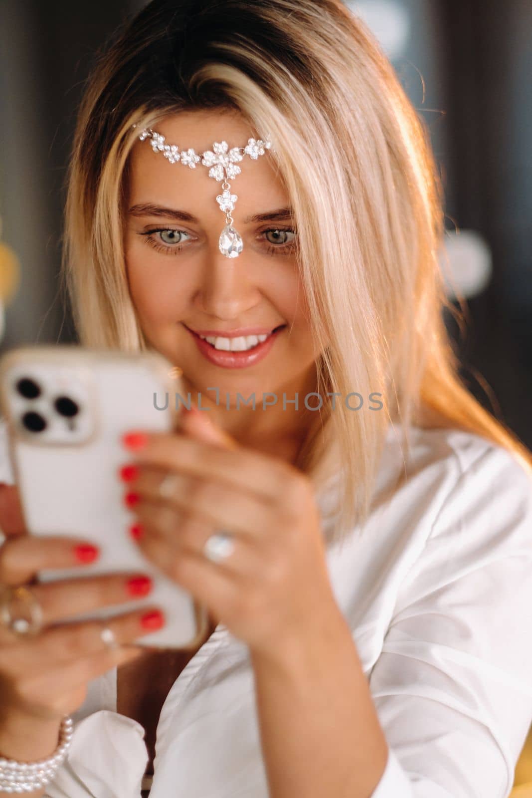 A positive young woman in a white dress and an ornament on her head was smiling, holding a phone in her hands and looking into it by Lobachad