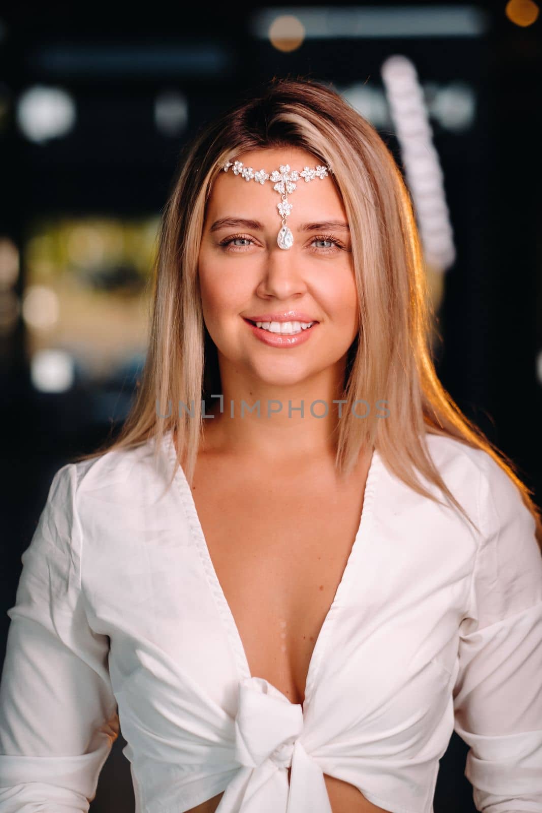 A positive young woman in a white dress and an ornament on her head smiles at the camera while standing in the studio by Lobachad