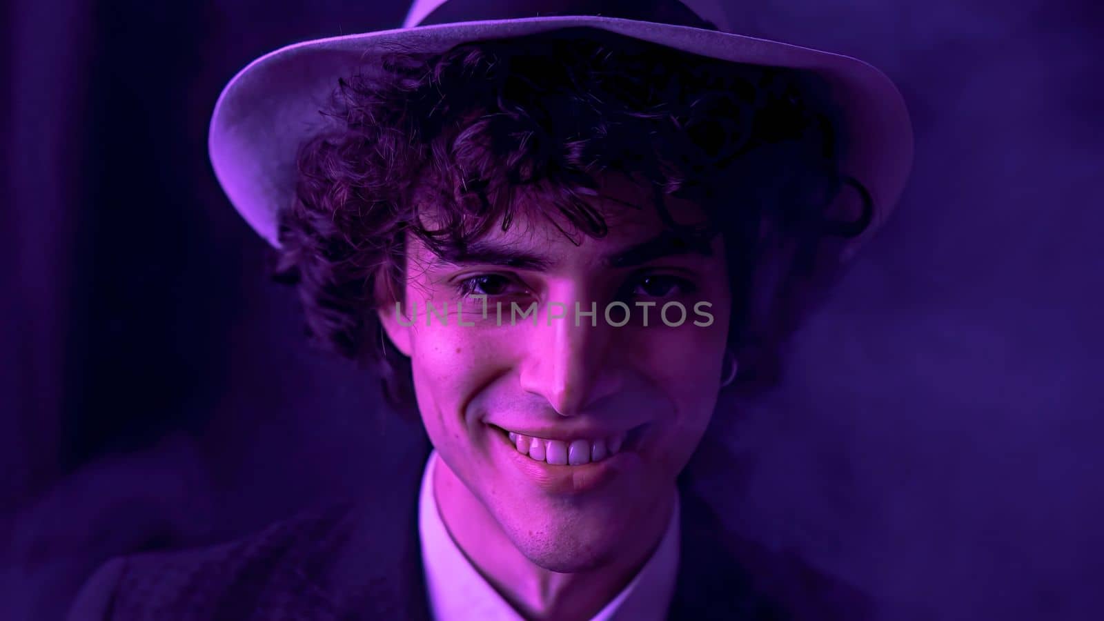 A young man with dark brown hair is happily singing and dancing with comical facial expressions in front of a smoky, dark background. He is wearing a stylish black suit and a fashionable hat.