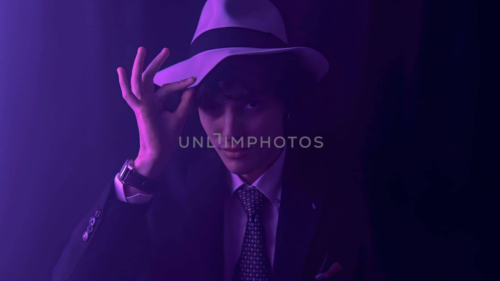 A handsome, curly haired man strikes a playful pose as he dances in a sleek black suit. Touching his a cowboy hat and a mischievous wink towards the camera. Against a dramatic, dark background.