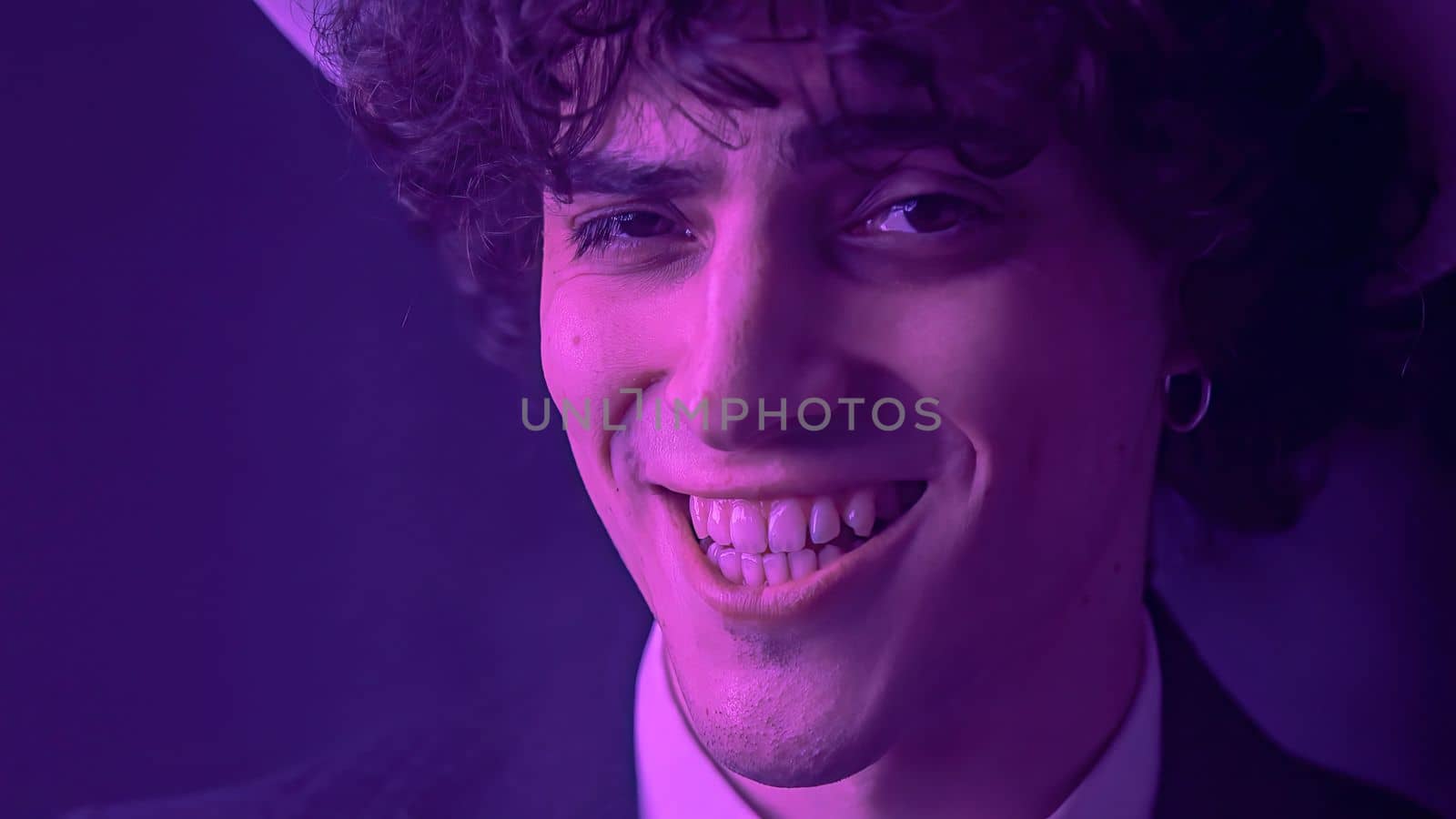 An attractive young man with messy hair and a chiseled jawline beams at the camera with a toothy smile. With a background of deep darkness, this handsome individual exudes charm and inviting expression.